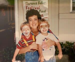 A woman holds two children with painted faces, smiling in front of a sign for a festival.